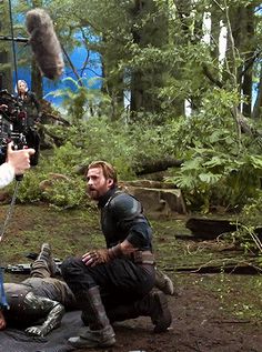 two men are filming in the woods with one man holding a camera and another person sitting on the ground