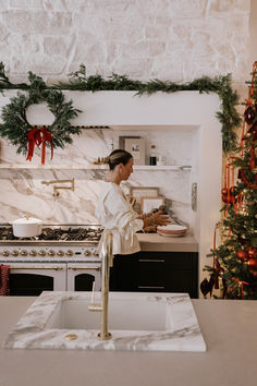 a woman standing in front of a kitchen counter next to a stove top oven and christmas wreath