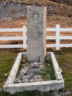 a grave in the middle of a field with a fence behind it and grass growing around it