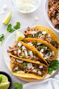 three shredded pork tacos on a white plate with limes and cilantro