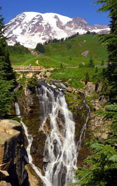 a large waterfall in the middle of a forest with snow covered mountains behind it and a bridge