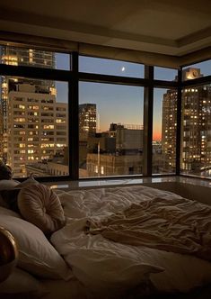 an unmade bed in front of a large window with the city lights out at night