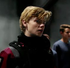 a young man with blonde hair wearing a red and black jacket looking at the camera