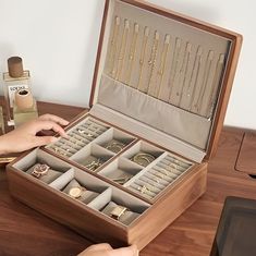 an open wooden jewelry box on a table next to a bottle of perfume and two women's hands