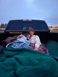 two people laying in the back of a pick up truck with a blanket on it