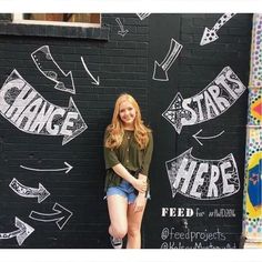 a woman standing in front of a black wall with white writing and arrows on it