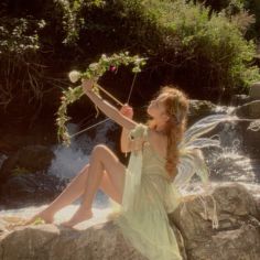 a beautiful young woman sitting on top of a rock next to a waterfall holding flowers