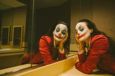 two women with clown makeup are looking at themselves in the mirror, one is holding her face up