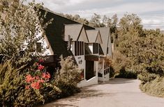 the house is surrounded by trees and bushes with red flowers on each side, along with a driveway leading up to it