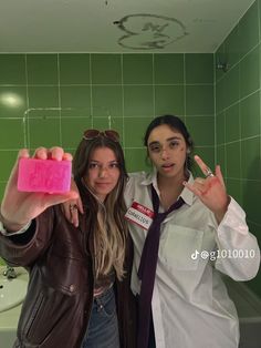 two people posing for the camera in a green tiled bathroom, one holding up a pink object