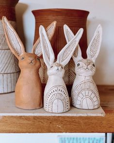 three ceramic rabbits sitting next to each other on top of a wooden shelf with vases in the background