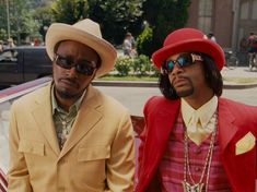 two men standing next to each other in front of a car wearing hats and sunglasses