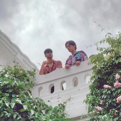 two men standing on a white fence with pink flowers in the foreground and dark clouds in the background