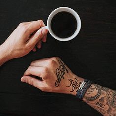 two people holding hands over a table with a cup of coffee and an airplane tattoo on their arm