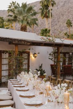 a long table set with white plates and gold place settings in front of palm trees