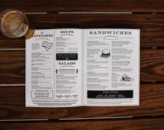 an open menu sitting on top of a wooden table next to a glass of beer