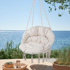 a white hanging chair next to a basket and some plants on the ground with water in the background