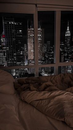 an unmade bed in front of a large window overlooking the city lights at night