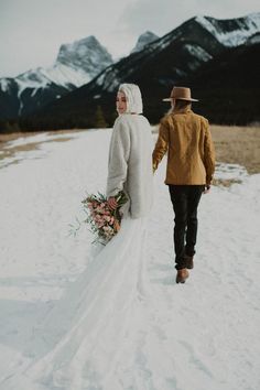 a bride and groom walking through the snow holding hands with mountains in the background on their wedding day