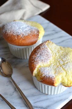 two muffins with powdered sugar sit on a marble plate next to a spoon