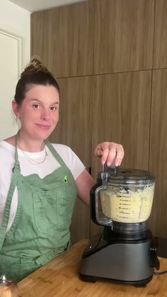 a woman standing in front of a blender on top of a wooden cutting board