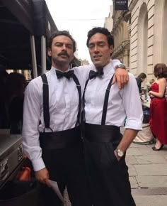 two men standing next to each other in front of a building wearing black suspenders and bow ties