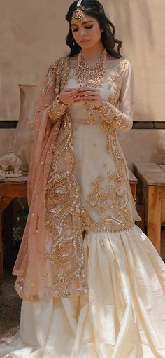 a woman in a white and gold wedding dress standing next to a bench with her hands together