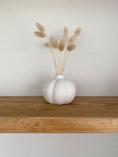 a white vase filled with dry grass on top of a wooden shelf