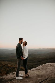 an engaged couple kissing on top of a rock in the mountains at sunset or sunrise