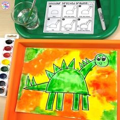 an orange tray with watercolors and a drawing of a green dinosaur