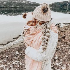 a woman holding a baby in her arms near the water with mountains in the background