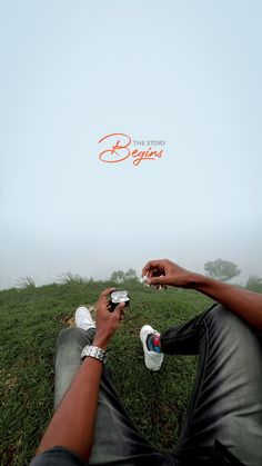 a man sitting on top of a lush green field holding onto his cell phone with the sky in the background