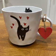 a white coffee cup with a black cat on it and a red heart hanging from the handle