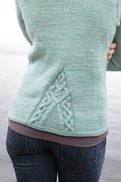 a woman wearing a green sweater with an intricate design on the back and sides, standing in front of a body of water