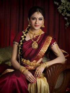 a woman in a red and gold sari sitting on a chair with her hands clasped to her chest