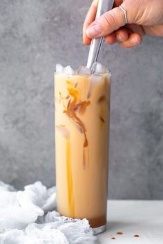 someone is holding a spoon in a tall glass filled with ice and caramel colored liquid