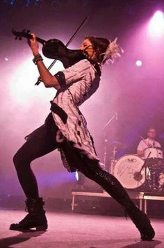 a man with long hair holding a violin on stage in front of purple lights and two drummers behind him