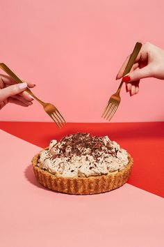 two hands holding forks over a pie on a pink and red surface with one hand reaching for it