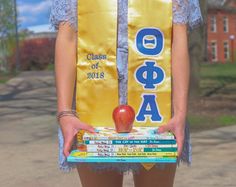 a woman holding a stack of books and an apple in her hands while wearing a graduation sash