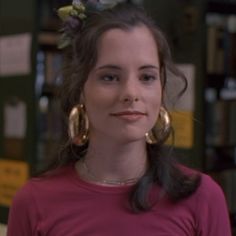 a woman wearing large gold hoop earrings and a pink shirt in front of bookshelves