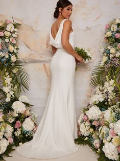 a woman standing in front of flowers and greenery wearing a white gown with one shoulder open