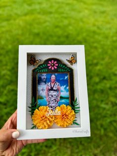 a person holding up a small photo frame with flowers in front of them and butterflies on the grass