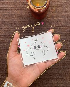 a person holding up a card with a drawing on it next to a cup of tea