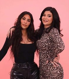 two women standing next to each other in front of a pink background and one is wearing black