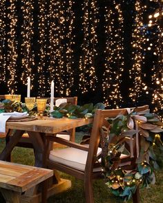 an outdoor table set for two with candles and greenery on the side, surrounded by lights in the background