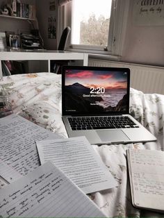 an open laptop computer sitting on top of a bed next to sheets of paper and notebooks