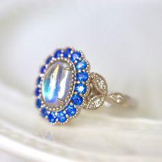 Mesmerizing Blue Moonstone Halo Ring. Moonstone engagement ring. Blue sapphires & diamonds. Edwardian Moonstone. 14K White Gold. US 7.25 A beautiful and unique moonstone ring, created with precision cut sapphires in 14K white gold, a luxurious and high-quality piece of jewelry, perfect to express your love. 💎 PREMIUM RING -- A brand new moonstone ring, personally designed and made with the highest quality of 14K white gold.  ✨ CLASSIC BRILLIANCE -- Designed with a beautiful blue moonstone at th Blue Oval Moonstone Ring Fine Jewelry, Anniversary Moonstone Ring With Blue Topaz Accent Stones, Anniversary Blue Topaz Moonstone Ring With Accent Stones, Blue Celestial Moonstone Ring, Heirloom Moonstone Ring With Accent Stones For Anniversary, Blue Moonstone Ring With Accent Stones, Celestial Blue Round Moonstone Ring, Celestial Blue Moonstone Ring, Celestial Sapphire Ring For Anniversary With Center Stone