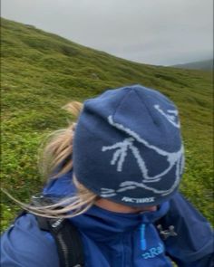 a person wearing a blue jacket and hat on top of a grass covered hill with hills in the background