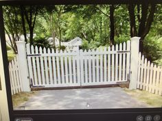 a white picket fence in the middle of a driveway with trees and grass around it