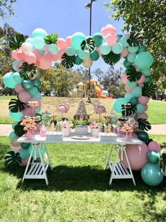 a table topped with balloons and desserts under an arch made out of palm leaves